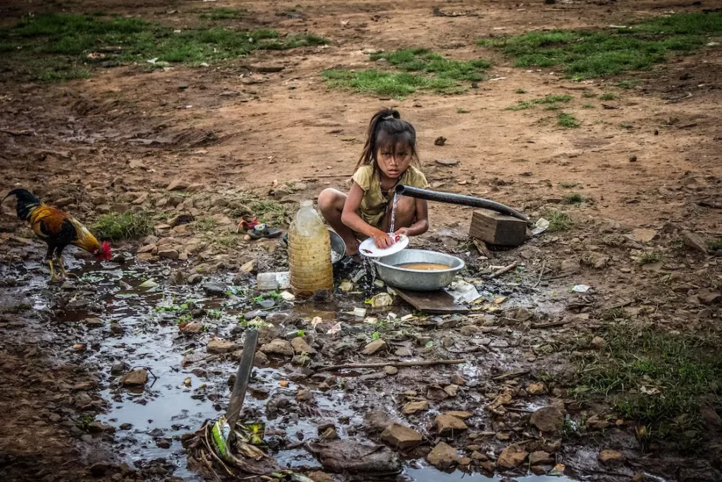 Girl washing dishes outside in a trickle of running water with mud and dirty water all around.