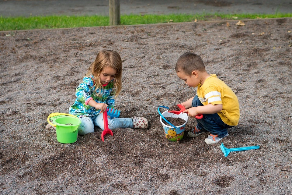 Boy and girl playing in the sand with buckets, toy rakes, and toy shovels.